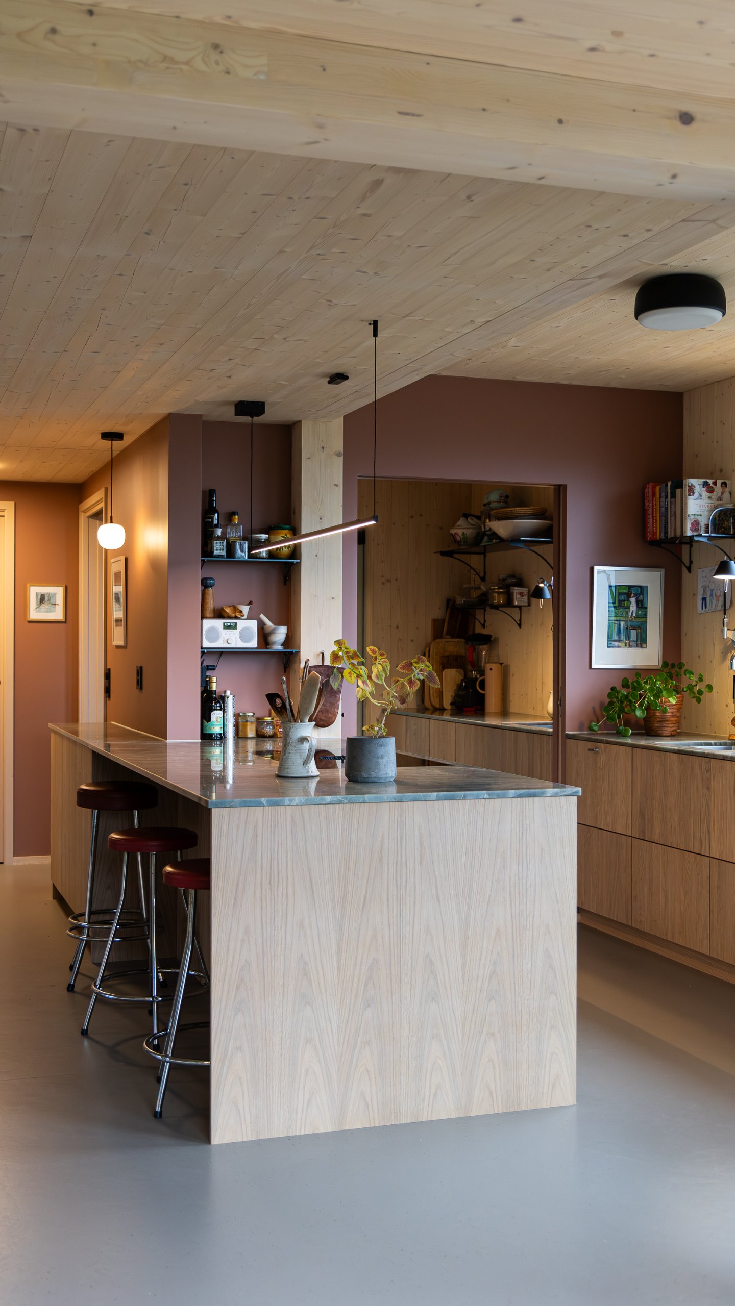 Köksö med tre barstolar under en smal pendellampa, omgiven av träpanel och öppna hyllor. Köksö med tre barstolar under en smal pendellampa, omgiven av träpanel och öppna hyllor.