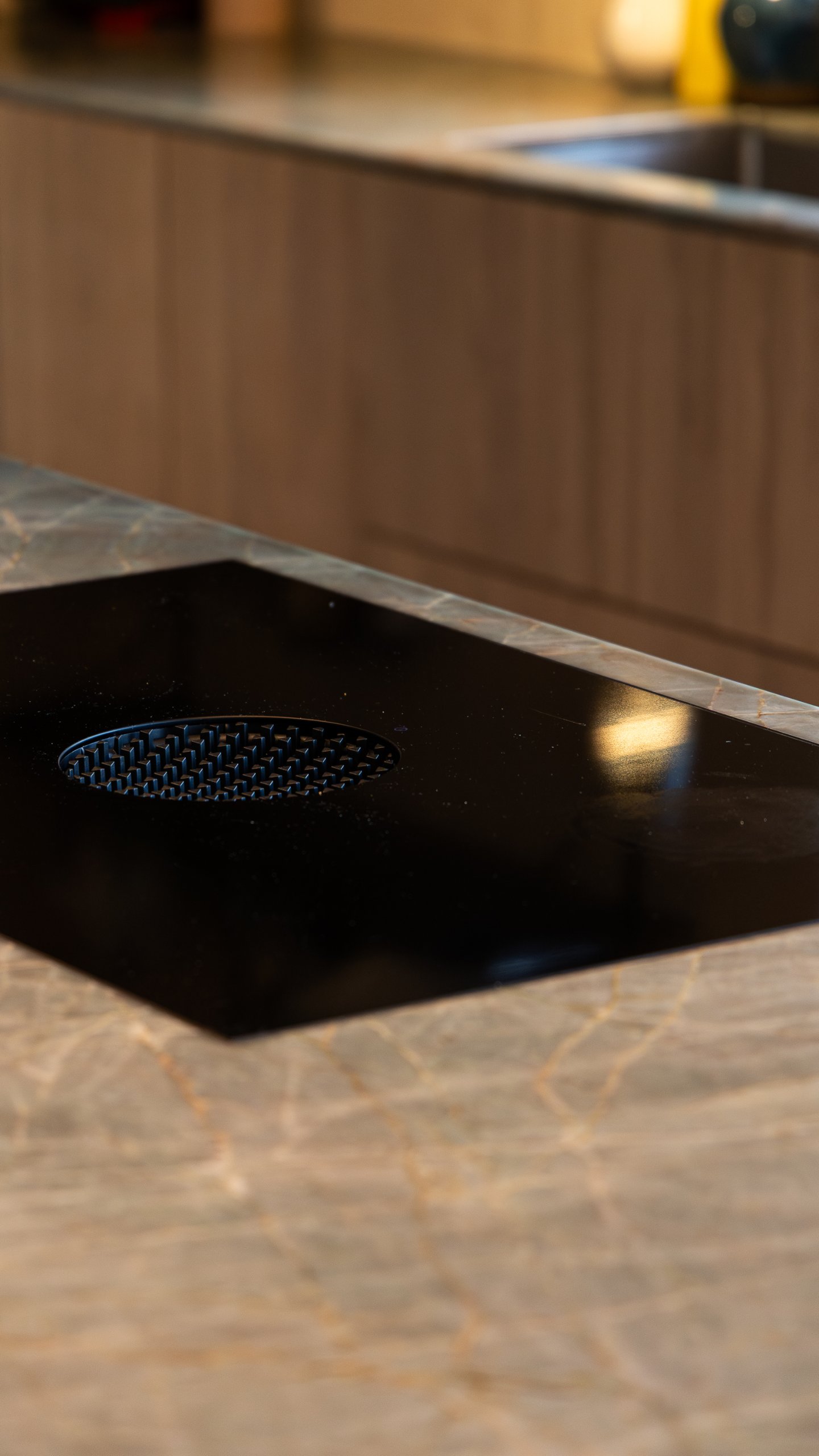 Närbild av svart häll med runt ventilationsgaller, infälld i köksöns ådrade stenskiva. Närbild av svart häll med runt ventilationsgaller, infälld i köksöns ådrade stenskiva.