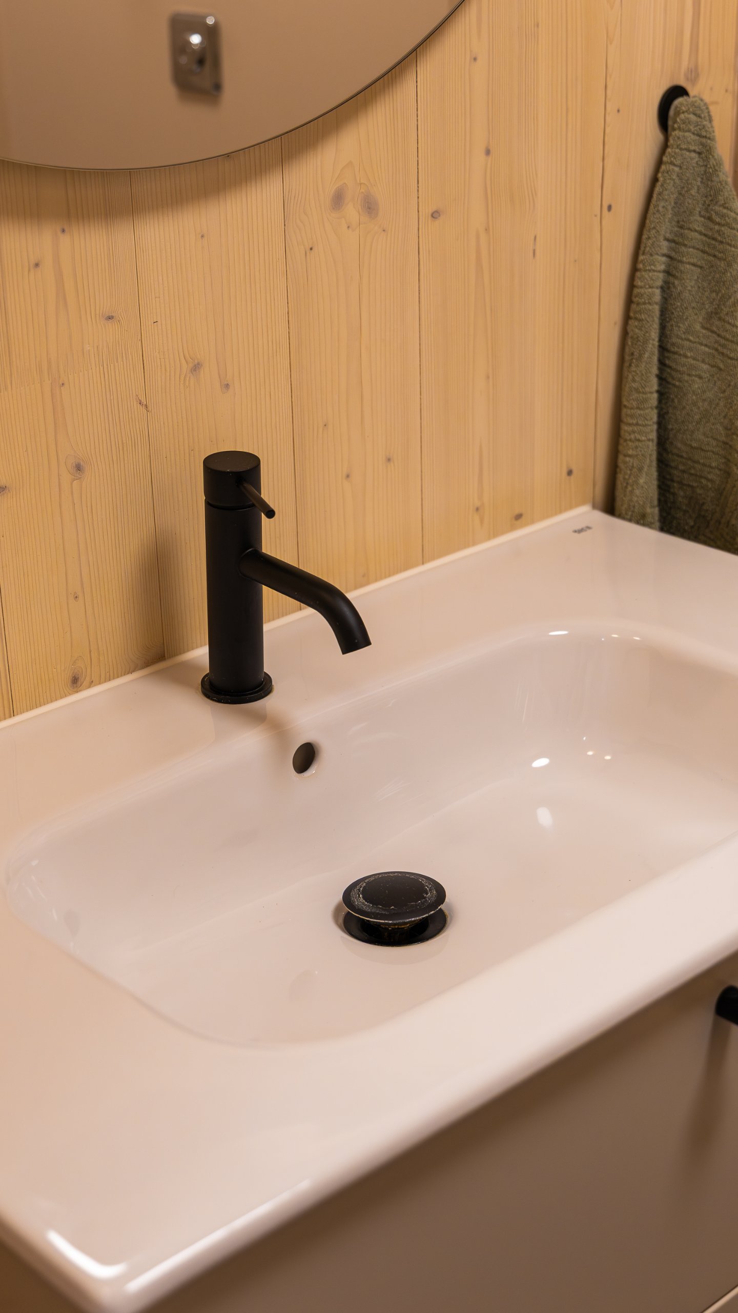 Vitt tvättställ med mattsvart blandare och svart bottenventil framför en träklädd vägg. Vitt tvättställ med mattsvart blandare och svart bottenventil framför en träklädd vägg.